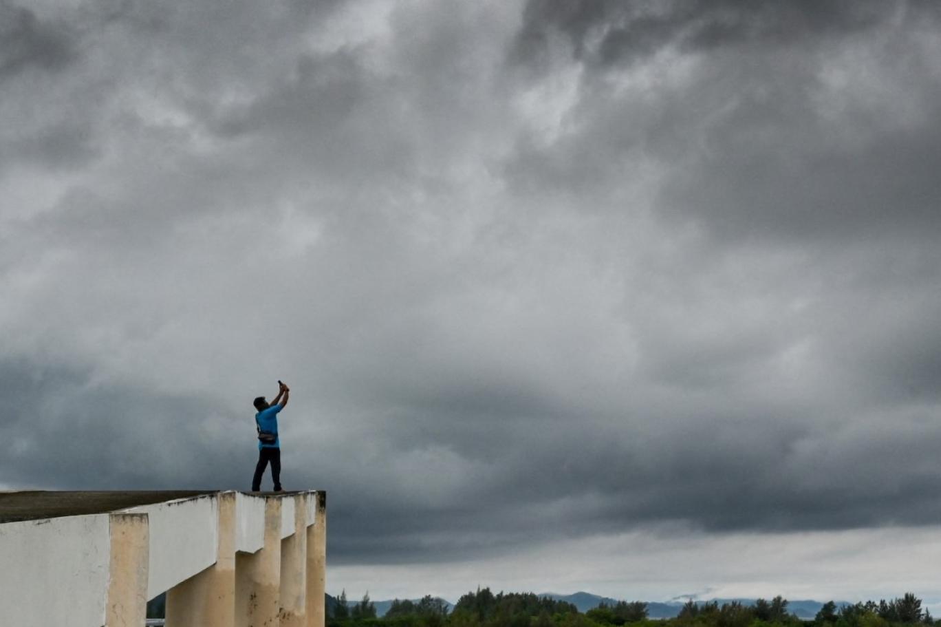 A silhouette of a man standing on top of a tsunami shelter building in Indonesia photographing dark storm clouds with a smartphone.